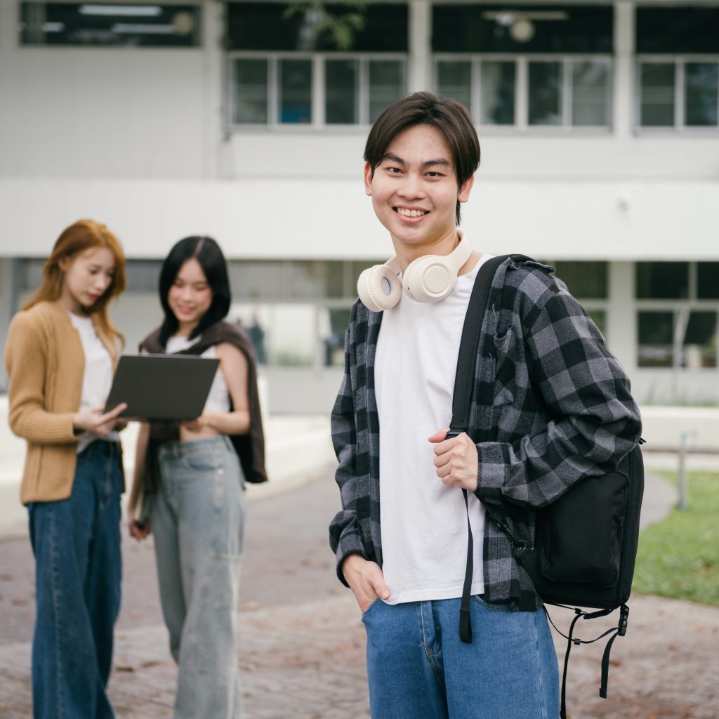 Happy male student standing on a university campus with headphones around his neck and a backpack, while two female students study on a laptop in the background