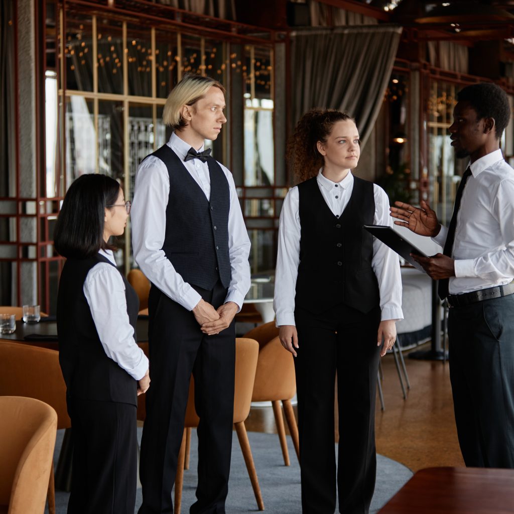 Staff meeting in luxury restaurant with Black young man as manager talking to servers wearing classic uniform